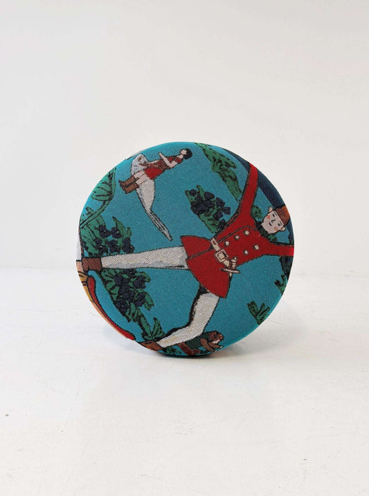 A cylindrical pouf covered in turquoise jacquard fabric, decorated with a naive-inspired figurative motif