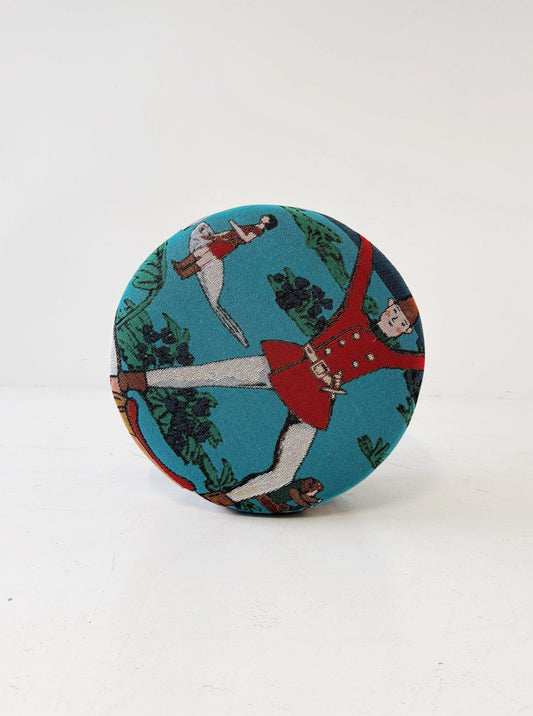 A cylindrical pouf covered in turquoise jacquard fabric, decorated with a naive-inspired figurative motif